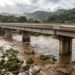 Ilustração de uma ponte rodoviária elevada de concreto sobre um rio com nível de água alto após chuvas intensas no Rio Grande do Sul.