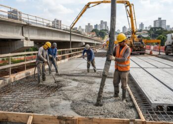 Trabalhadores com equipamentos de proteção acompanham o lançamento de concreto fresco por uma bomba-lança sobre as armaduras de um viaduto.