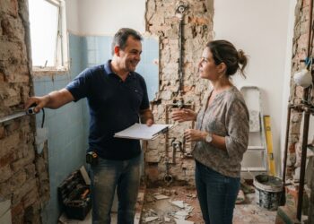 Profissional da construção mede um banheiro em reforma enquanto conversa com o proprietário sobre os serviços.
