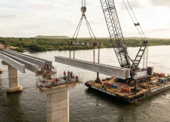 Representação de um guindaste instalado sobre uma balsa posicionando uma grande viga pré-moldada de concreto entre pilares de uma ponte no Rio São Francisco.