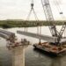 Representação de um guindaste instalado sobre uma balsa posicionando uma grande viga pré-moldada de concreto entre pilares de uma ponte no Rio São Francisco.
