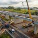 Representação de viaduto de concreto em construção na BR-116, com guindaste içando uma viga pré-moldada e caminhões circulando em pista separada.