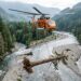 Helicóptero de carga transportando um grande tronco sobre um rio cercado por florestas durante uma operação de restauração ambiental.