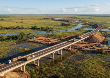 Representação aérea de uma rodovia com pontes de concreto atravessando áreas alagáveis do Pantanal, com caminhões pesados circulando sobre a pista elevada.