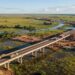 Representação aérea de uma rodovia com pontes de concreto atravessando áreas alagáveis do Pantanal, com caminhões pesados circulando sobre a pista elevada.