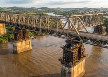Reconstrução ilustrativa de uma grande ponte metálica elevada sobre pilares de concreto por macacos hidráulicos, acima das águas do Rio Jacuí.