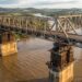 Reconstrução ilustrativa de uma grande ponte metálica elevada sobre pilares de concreto por macacos hidráulicos, acima das águas do Rio Jacuí.