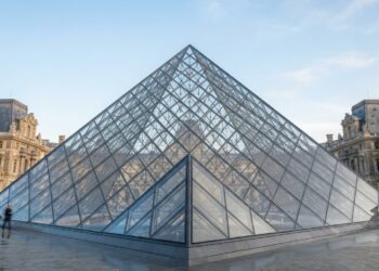 Pirâmide de vidro do Louvre com estrutura metálica aparente e o palácio histórico ao fundo.