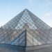 Pirâmide de vidro do Louvre com estrutura metálica aparente e o palácio histórico ao fundo.