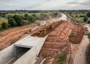 Vista aérea de um túnel ferroviário de concreto parcialmente coberto por terra, com escavadeiras e máquinas de terraplenagem trabalhando ao redor.