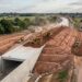 Vista aérea de um túnel ferroviário de concreto parcialmente coberto por terra, com escavadeiras e máquinas de terraplenagem trabalhando ao redor.