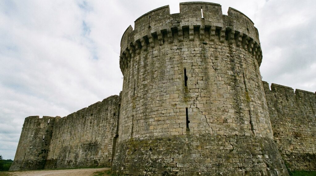Torre cilíndrica de pedra com seteiras e ameias em uma fortaleza medieval europeia.