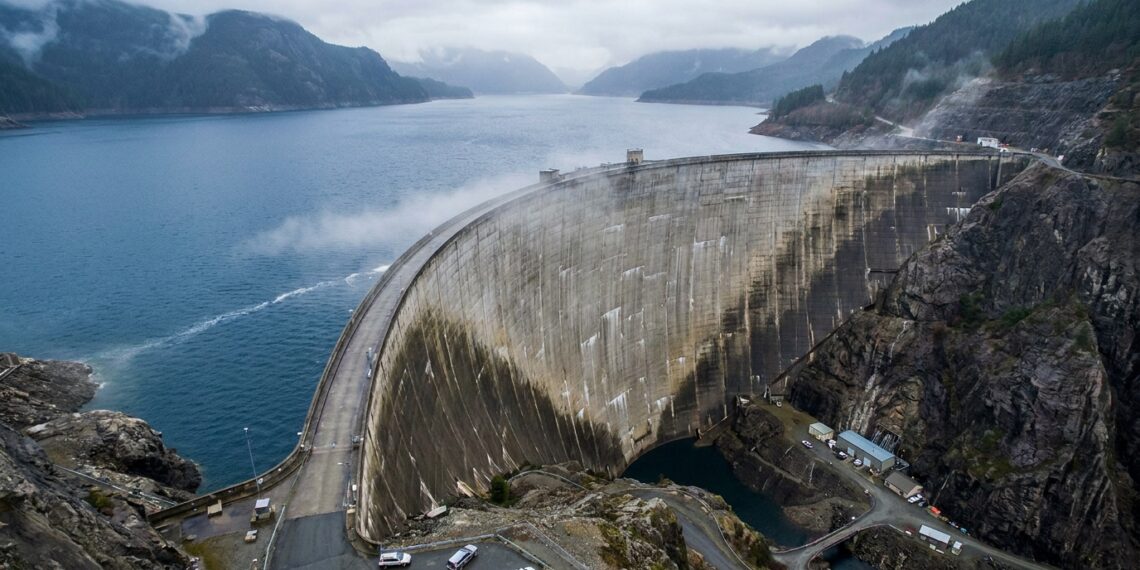 Barragem gigante de concreto contendo um enorme reservatório de água entre montanhas rochosas.