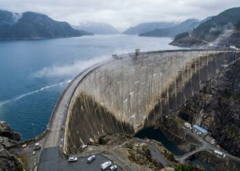 Barragem gigante de concreto contendo um enorme reservatório de água entre montanhas rochosas.