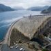 Barragem gigante de concreto contendo um enorme reservatório de água entre montanhas rochosas.