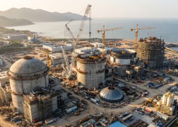 Vista aérea ilustrativa de uma usina nuclear chinesa em construção, com grandes edifícios de contenção de concreto e guindastes industriais.
