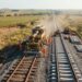 Vista aérea de uma representação da modernização da Malha Oeste, com máquinas ferroviárias, novos dormentes de concreto e trilhos em uma paisagem rural brasileira.