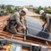 Técnicos com equipamentos de segurança instalam painéis solares na estrutura do telhado de uma residência.