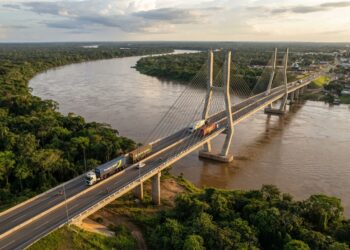 Representação da ponte binacional sobre o Rio Mamoré, com estrutura estaiada, caminhões e acessos rodoviários entre Brasil e Bolívia.