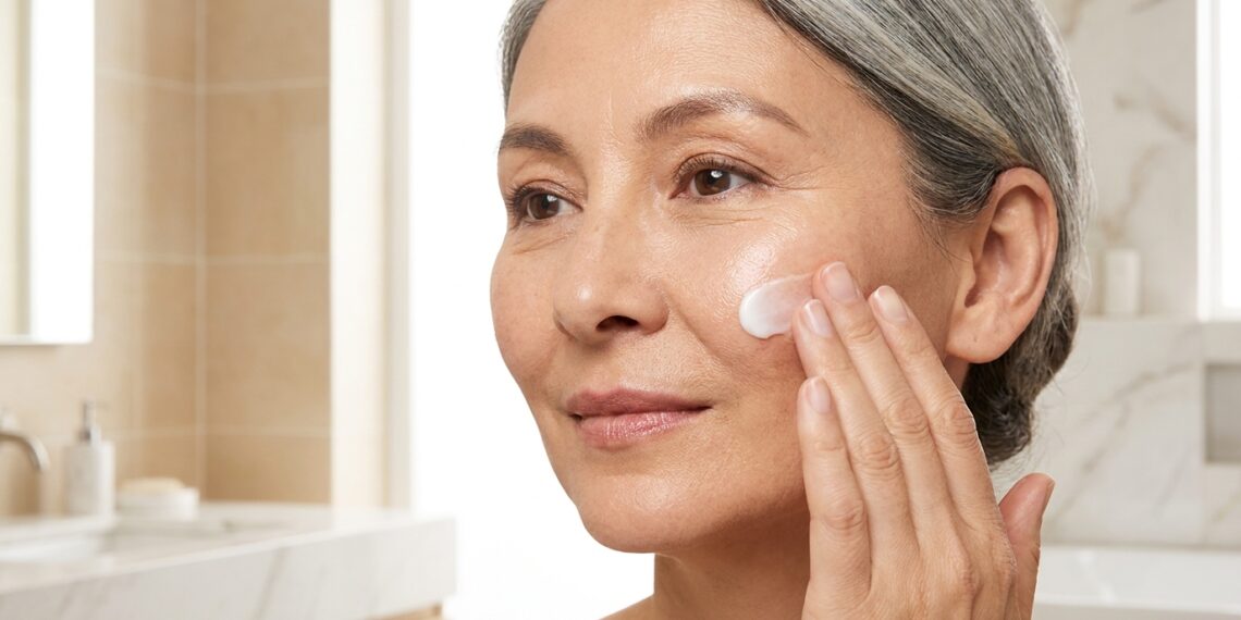 Mulher com cerca de 60 anos observa a textura da pele do rosto diante do espelho.