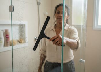 Mulher brasileira madura passa um rodinho de borracha no vidro molhado do box, retirando gotas de água após o banho.