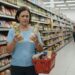 Mulher madura compara dois produtos diante de uma prateleira com muitas opções em um supermercado desconhecido.
