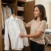 Mulher brasileira segura uma camisa branca recém-passada diante de um guarda-roupa aberto, verificando a peça antes de guardá-la.