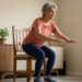 Mulher brasileira de aproximadamente 69 anos realiza exercício de sentar e levantar de uma cadeira para fortalecer as pernas.