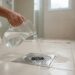 Mulher brasileira despeja água limpa de uma jarra transparente em ralo de aço inoxidável no piso de um banheiro limpo.