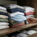 Mão retira uma camiseta do meio de uma pilha de roupas dobradas em um guarda-roupa, fazendo as peças superiores saírem do lugar.