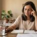 Mulher brasileira de aproximadamente 56 anos com expressão de cansaço mental, sentada à mesa com um caderno e um copo de água.