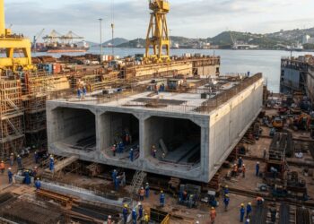 Representação de um enorme módulo retangular de concreto para túnel imerso em construção dentro de um dique seco, cercado por guindastes e trabalhadores.