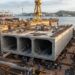 Representação de um enorme módulo retangular de concreto para túnel imerso em construção dentro de um dique seco, cercado por guindastes e trabalhadores.