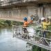 Técnicos realizam reparos localizados em uma viga de concreto sob uma ponte rodoviária sobre o Rio Doce.